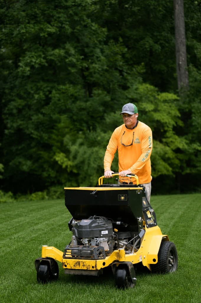 Simply Turf technician operating a stand-on Stinger aerator with seed box on a freshly striped lawn